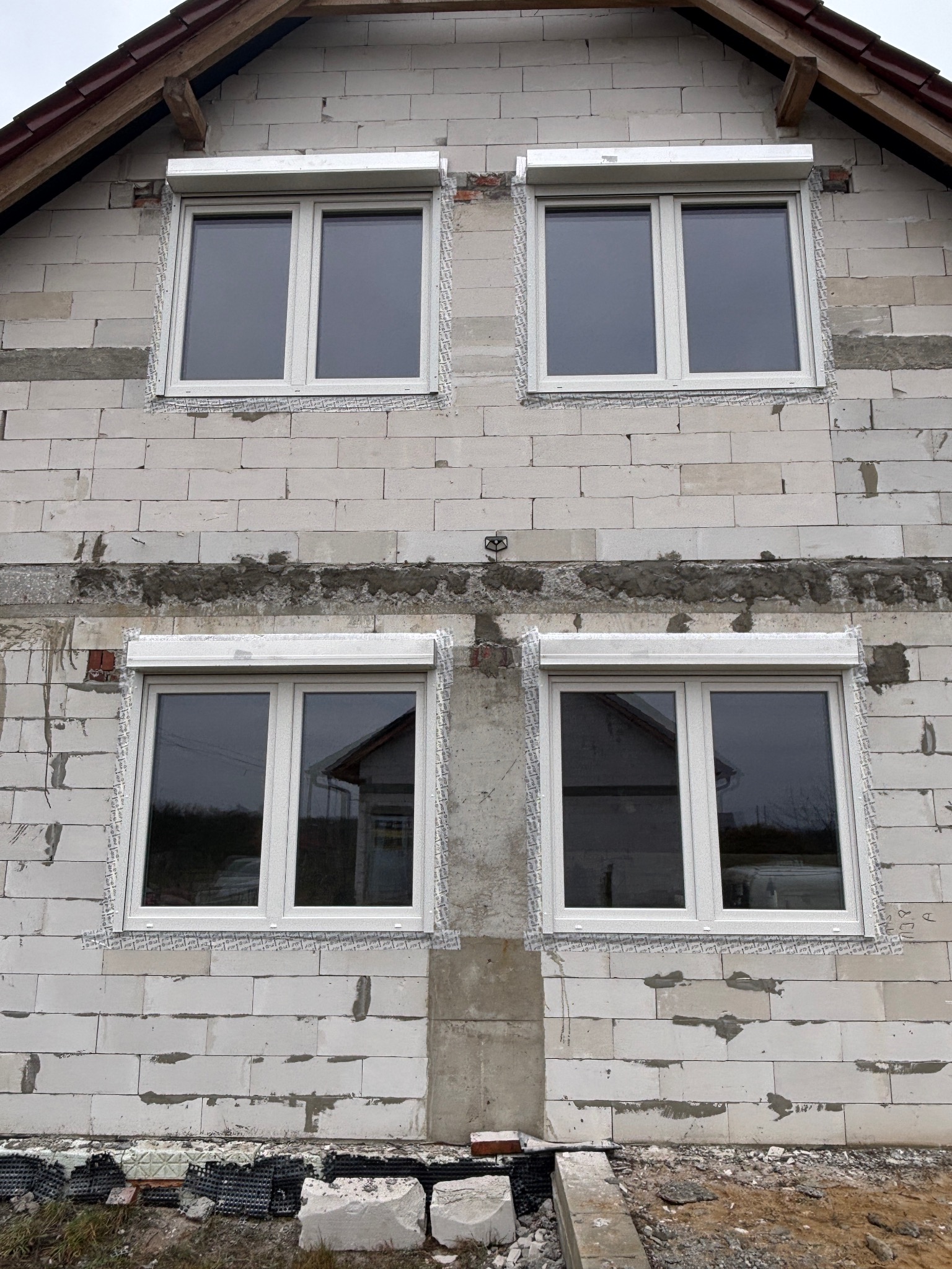 Montaż białych okien z roletami zewnętrznymi w surowym budynku z białego bloczka. Widoczne zabezpieczenia okien folią. Dom w trakcie budowy.