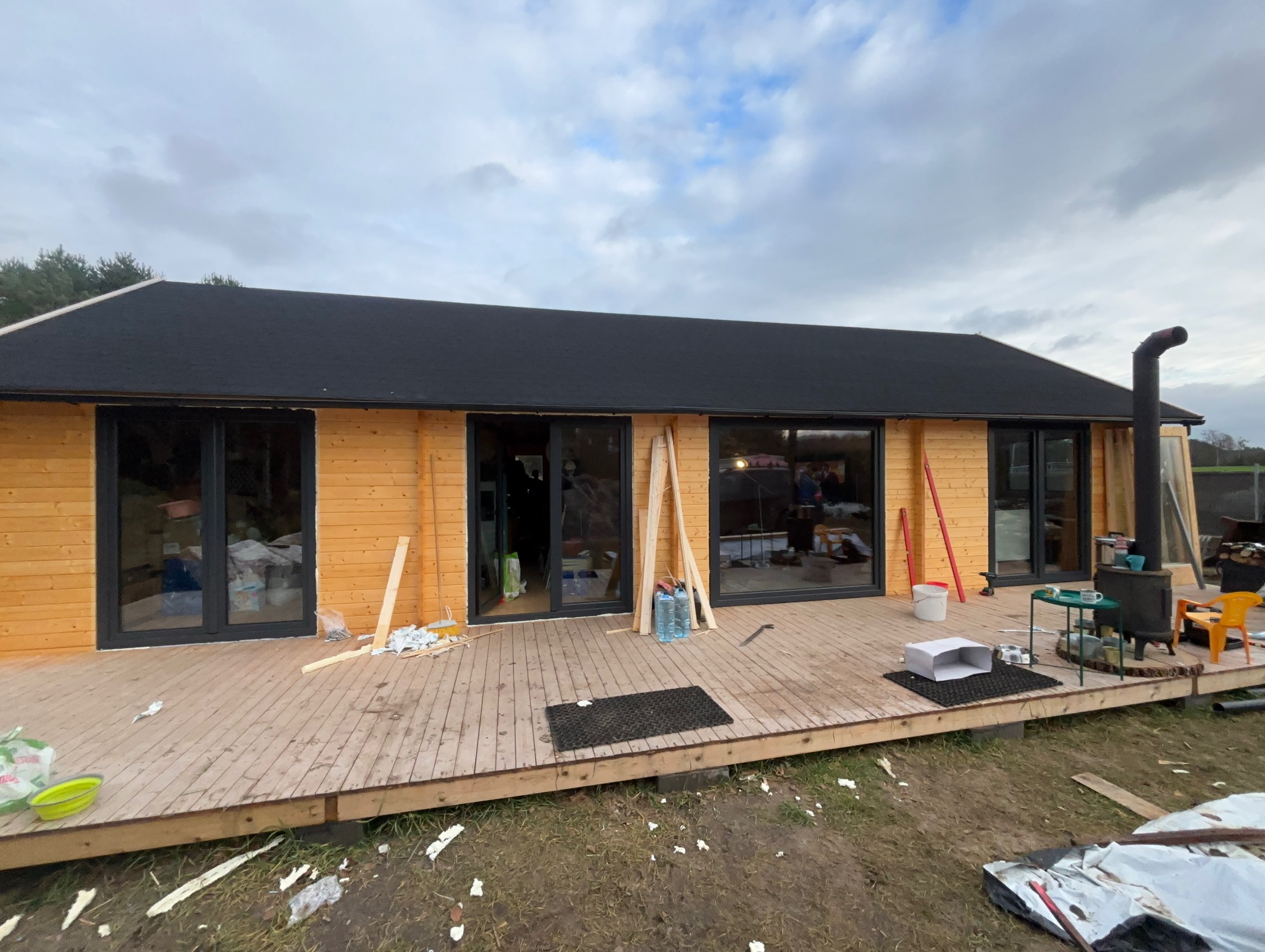 Drewniany dom z czarnymi ramami okiennymi i przeszklonymi drzwiami tarasowymi, widok z zewnątrz na drewniany taras z elementami wyposażenia i narzędziami.
