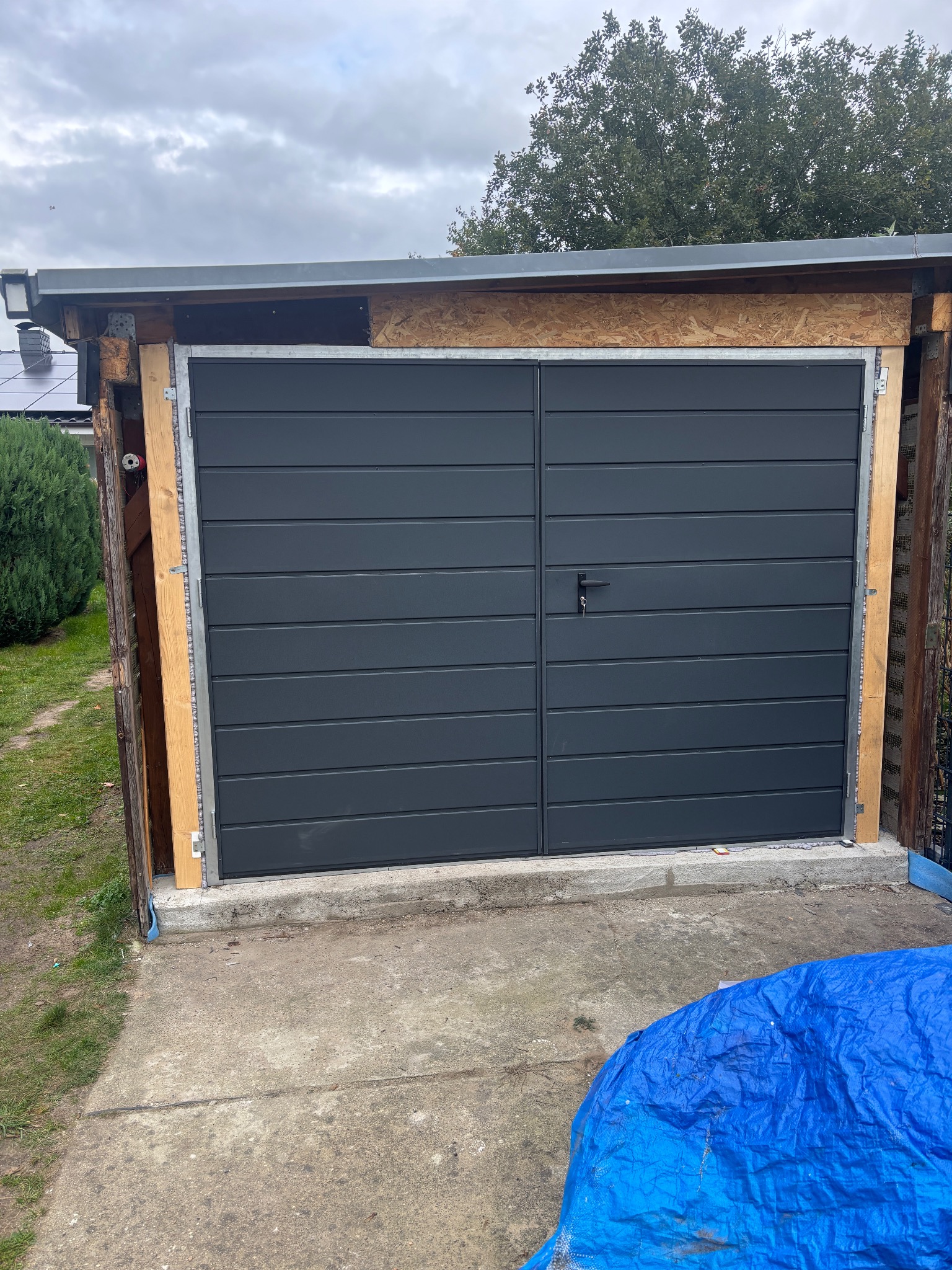 Szara, dwuskrzydłowa brama garażowa z poziomymi panelami, zamontowana w drewnianej konstrukcji. Na dole widoczna betonowa podstawa i fragment niebieskiej plandeki.