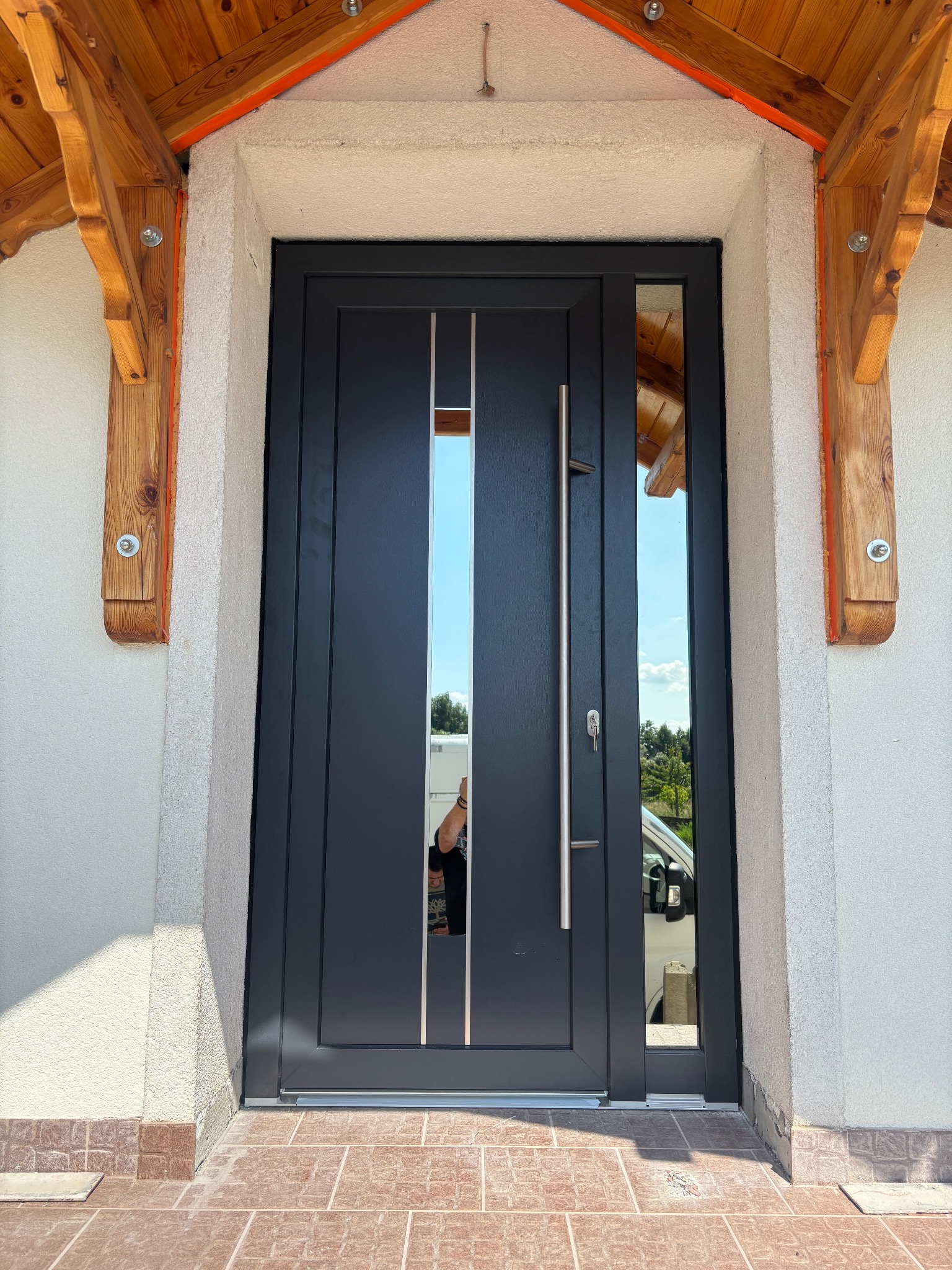 Grafitowe drzwi wejściowe z przeszkleniem i długim uchwytem, zamontowane w jasnej elewacji z drewnianym zadaszeniem. Widoczne odbicie otoczenia w szybie.