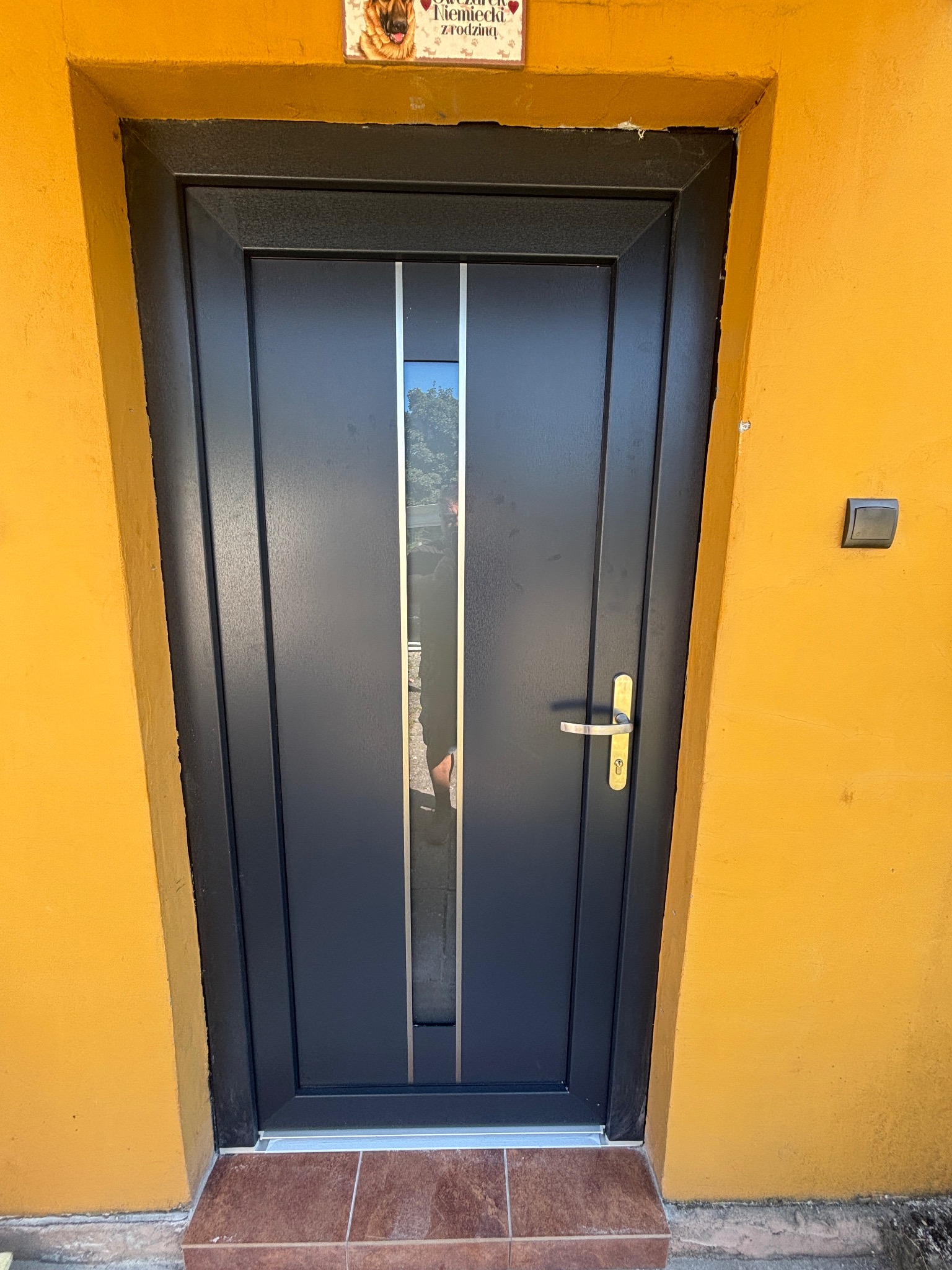 Ciemnoszare drzwi wejściowe z przeszkleniami i złotą klamką, na tle pomarańczowej ściany. Nad drzwiami tabliczka z owczarkiem niemieckim. Widoczny fragment brązowych płytek.