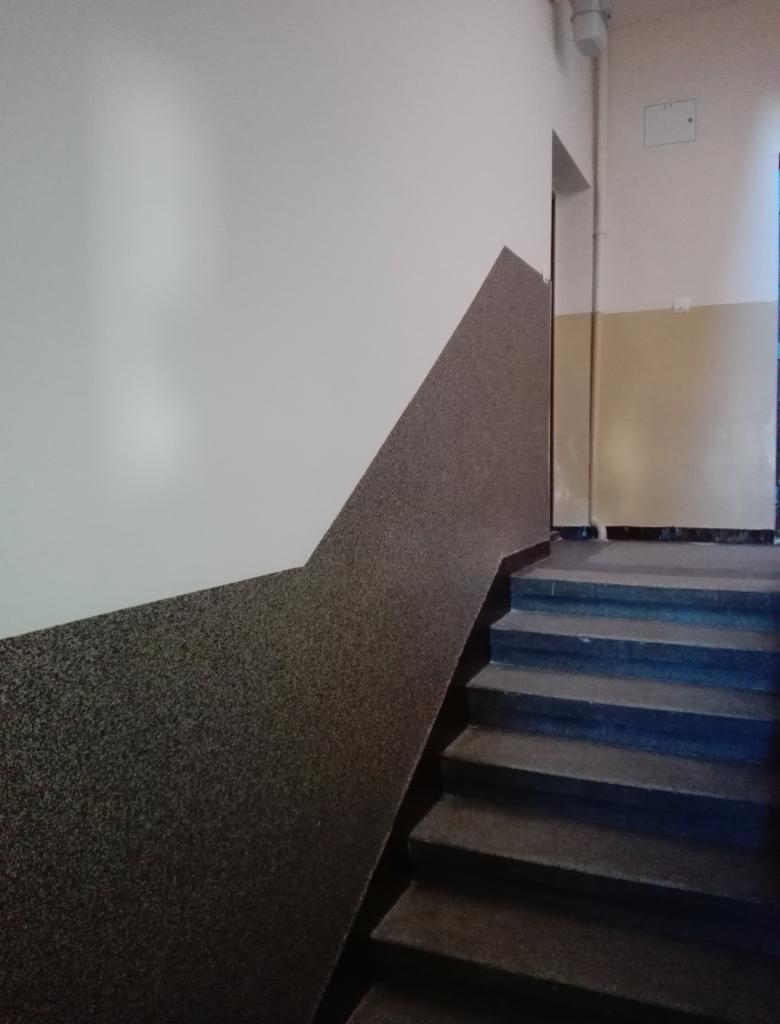 Klatka schodowa z wykończoną ścianą: dolna część pokryta szorstką, ciemną strukturą w kształcie trójkąta, górna część biała, gładka. Widoczne schody z ciemnego kamienia.