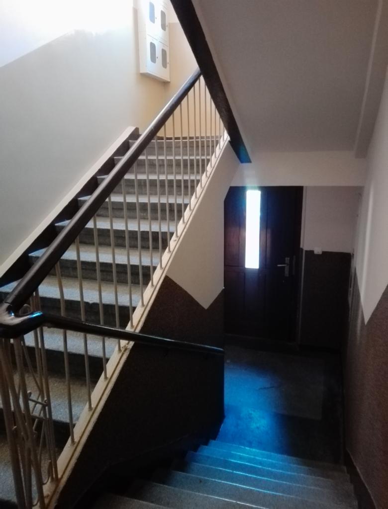 Wnętrze klatki schodowej w bloku z szarymi schodami, metalową balustradą i brązowymi poręczami. Ściany pomalowane na kremowo z brązowym pasem na dole. Na ścianie skrzynka elektryczna. Na końcu...