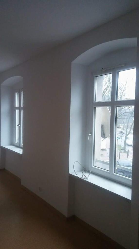 Wnętrze pokoju z dwoma oknami o łukowatych nadprożach po malowaniu, widoczne niedoskonałości w wykończeniu przy oknach i kabel zwisający na parapecie.