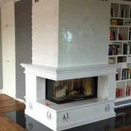 Biały, kaflowy kominek z dekoracyjnymi zdobieniami w rogu pokoju, z widocznym paleniskiem i czarną wkładką, sąsiadujący z zabudowaną szafą i regałem z książkami.