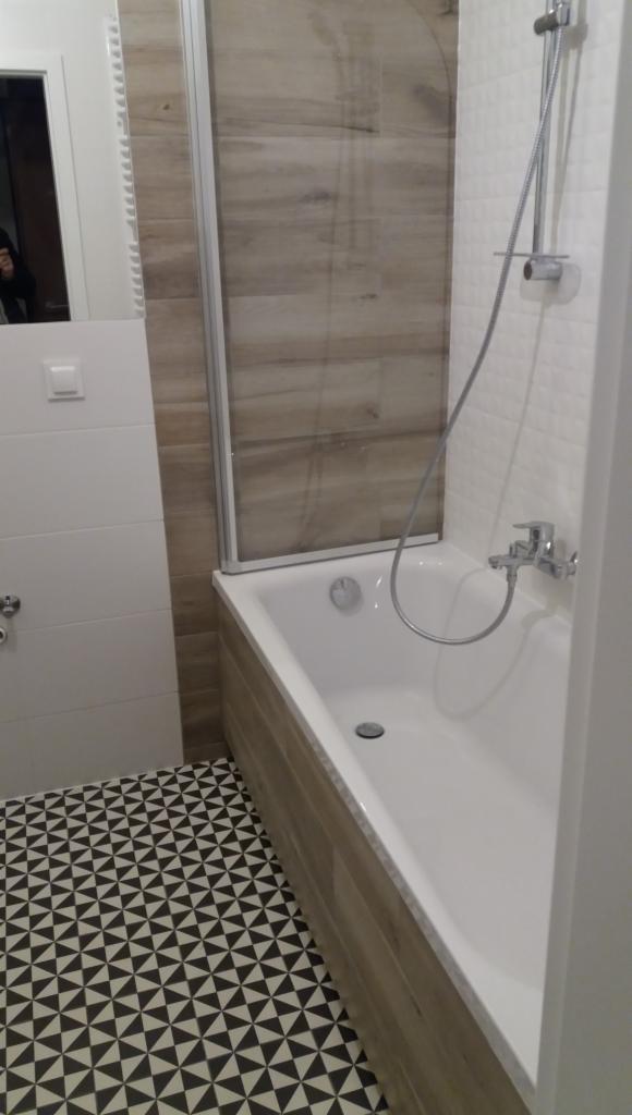 Biała wanna z parawanem nawannowym i baterią prysznicową, obudowana płytkami imitującymi drewno, z podłogą w czarno-białe trójkąty.