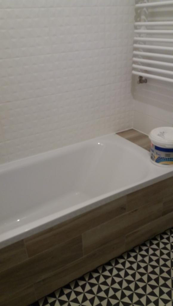 Wykończona łazienka z białą wanną obudowaną panelami imitującymi drewno, białe płytki 3D na ścianie, grzejnik drabinkowy i wzorzysta podłoga w czarno-białe trójkąty. Na wannie stoi wiaderko z farbą.