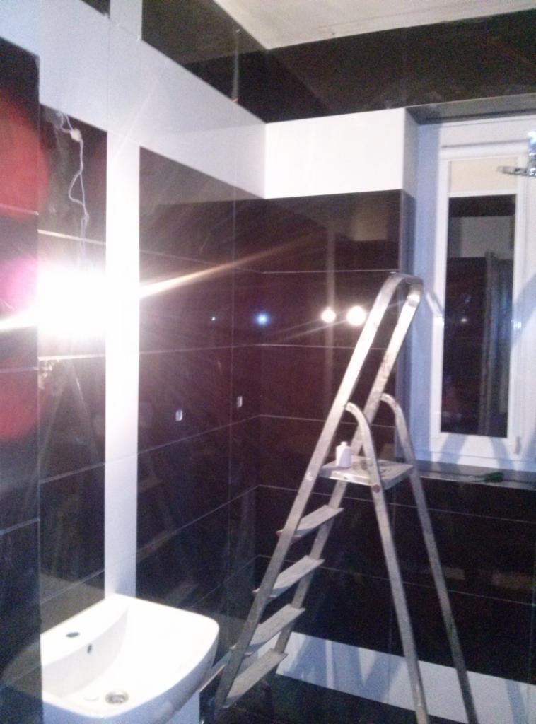 Łazienka w trakcie remontu: ciemne, błyszczące płytki, białe fugi i listwy, aluminiowa drabina z pojemnikiem na farbę, umywalka w narożniku, widoczne okno.