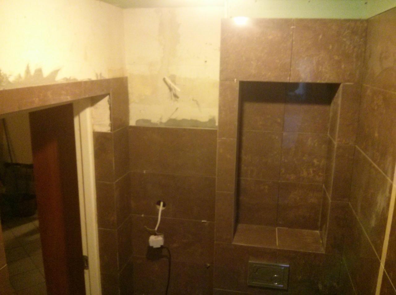 Łazienka w trakcie remontu, widoczne brązowe płytki, wnęka w ścianie wyłożona płytkami, niedokończone ściany i wystające kable elektryczne.