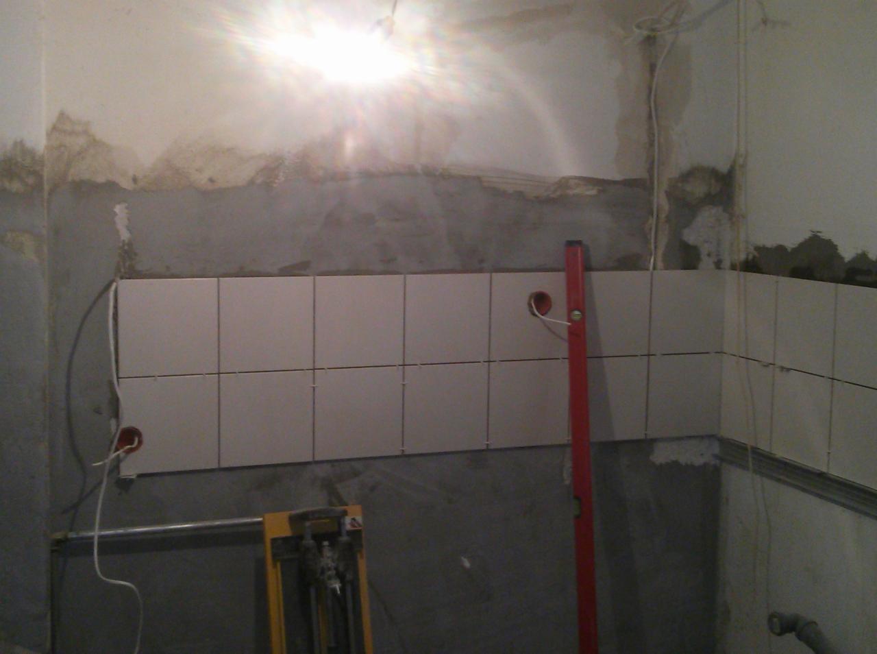 Ściana w trakcie układania jasnych, kwadratowych płytek, widoczny poziomica, narzędzie do cięcia płytek, oraz niedokończone ściany z przewodami elektrycznymi wystającymi ze ściany.