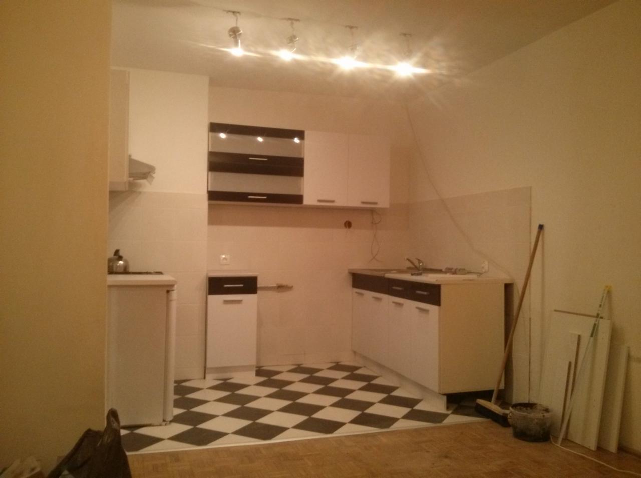Kuchnia po remoncie z biało-czarną szachownicą na podłodze, białe szafki z ciemnymi blatami i elementami, widoczne ślady po remoncie, w rogu oparta miotła i płyty.