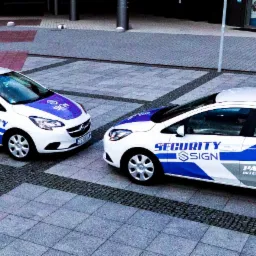 Dwa białe samochody patrolowe z niebieskimi akcentami i logo firmy Security Sign zaparkowane na placu z kostki brukowej.