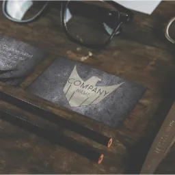 Dwie wizytówki z logo orła na szorstkim, ciemnym tle, otoczone okularami, ołówkami i nożyczkami na drewnianym stole.