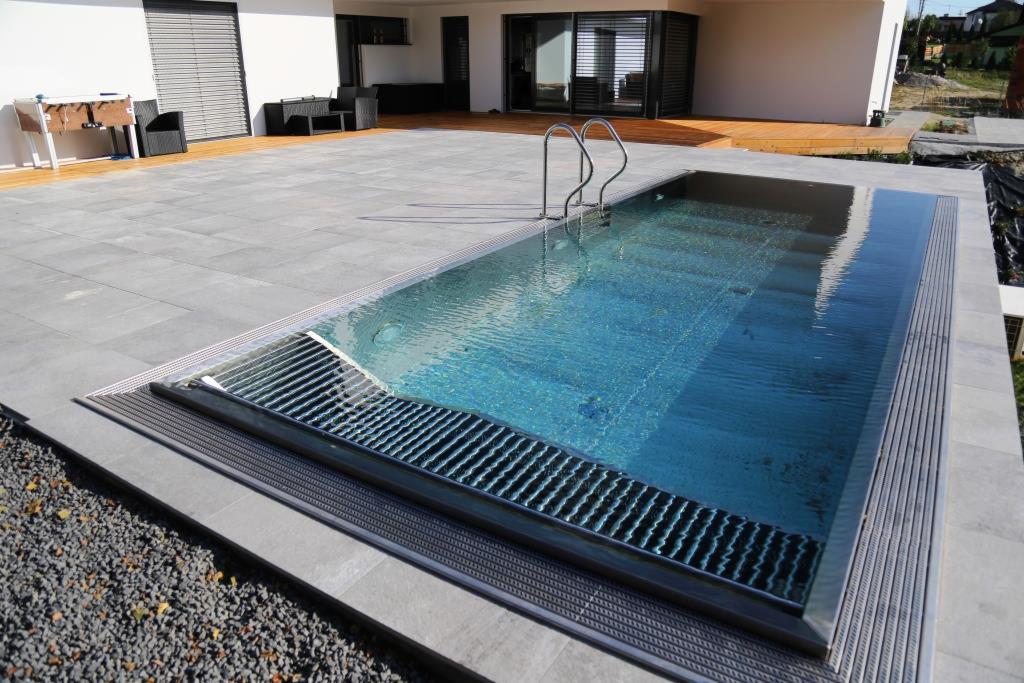 Nowoczesny basen bez krawędzi z metalową kratką odpływową i schodami, otoczony szarymi płytami tarasowymi i drewnianym podestem, na tle minimalistycznego domu z dużymi oknami.