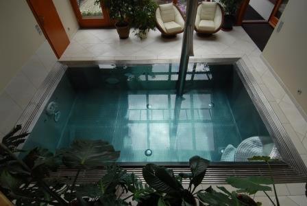 Wewnętrzny basen z turkusową wodą i dyszami, widok z góry, z roślinami w pierwszym planie i dwoma fotelami w tle.