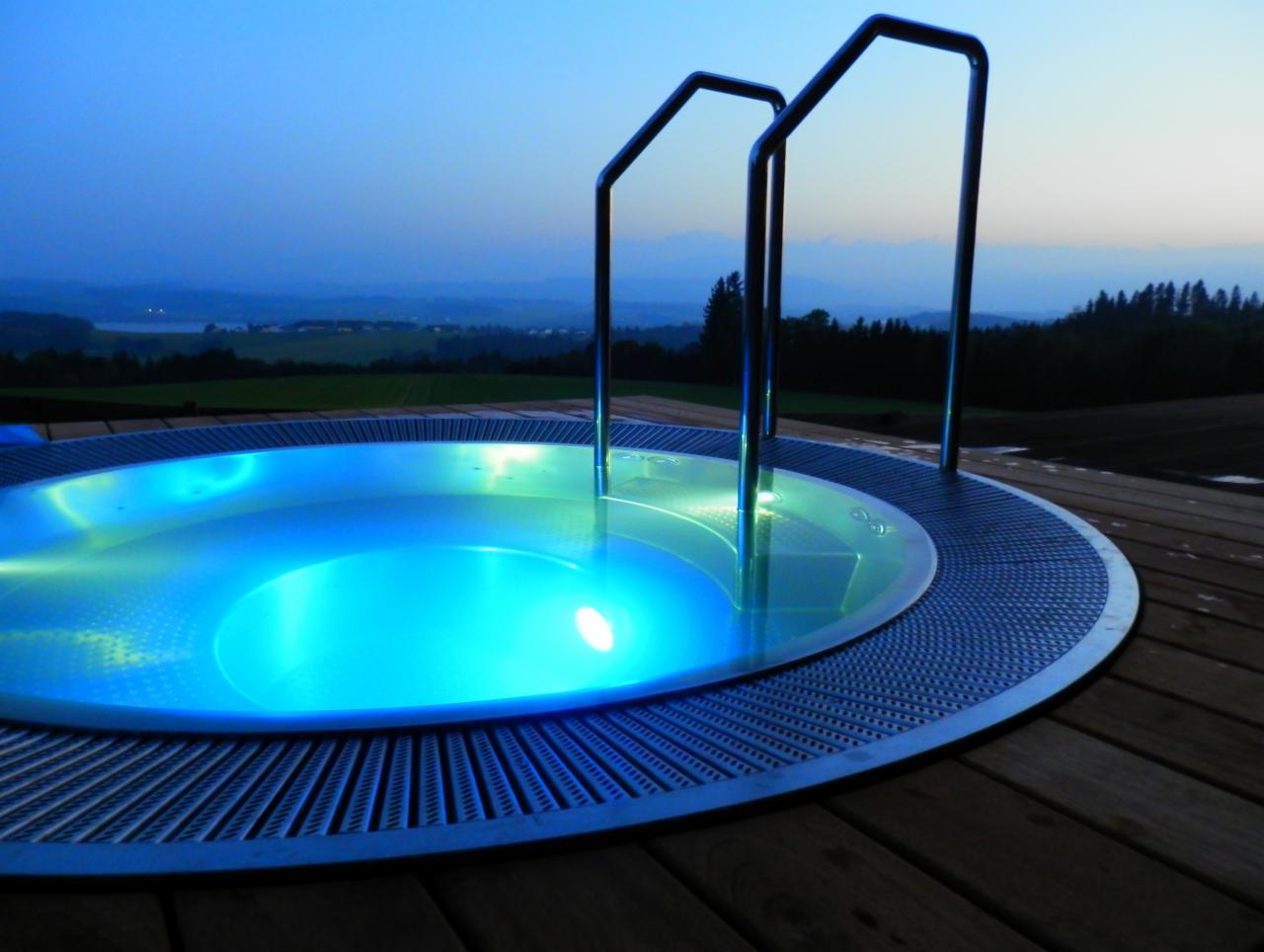 Okrągły, podświetlany na niebiesko basen na drewnianym tarasie z metalowymi schodkami, w tle krajobraz z lasem i zachodzącym słońcem.