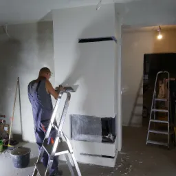 Pracownik na drabinie wygładza szpachelką ścianę obudowy wkładu kominkowego, wzmocnioną siatką, w surowym wnętrzu.