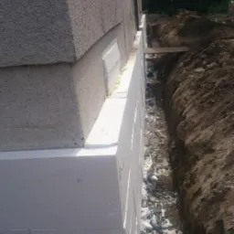 Ocieplenie fundamentów budynku styropianem, widoczny wykop i fragment elewacji z szorstkim tynkiem.