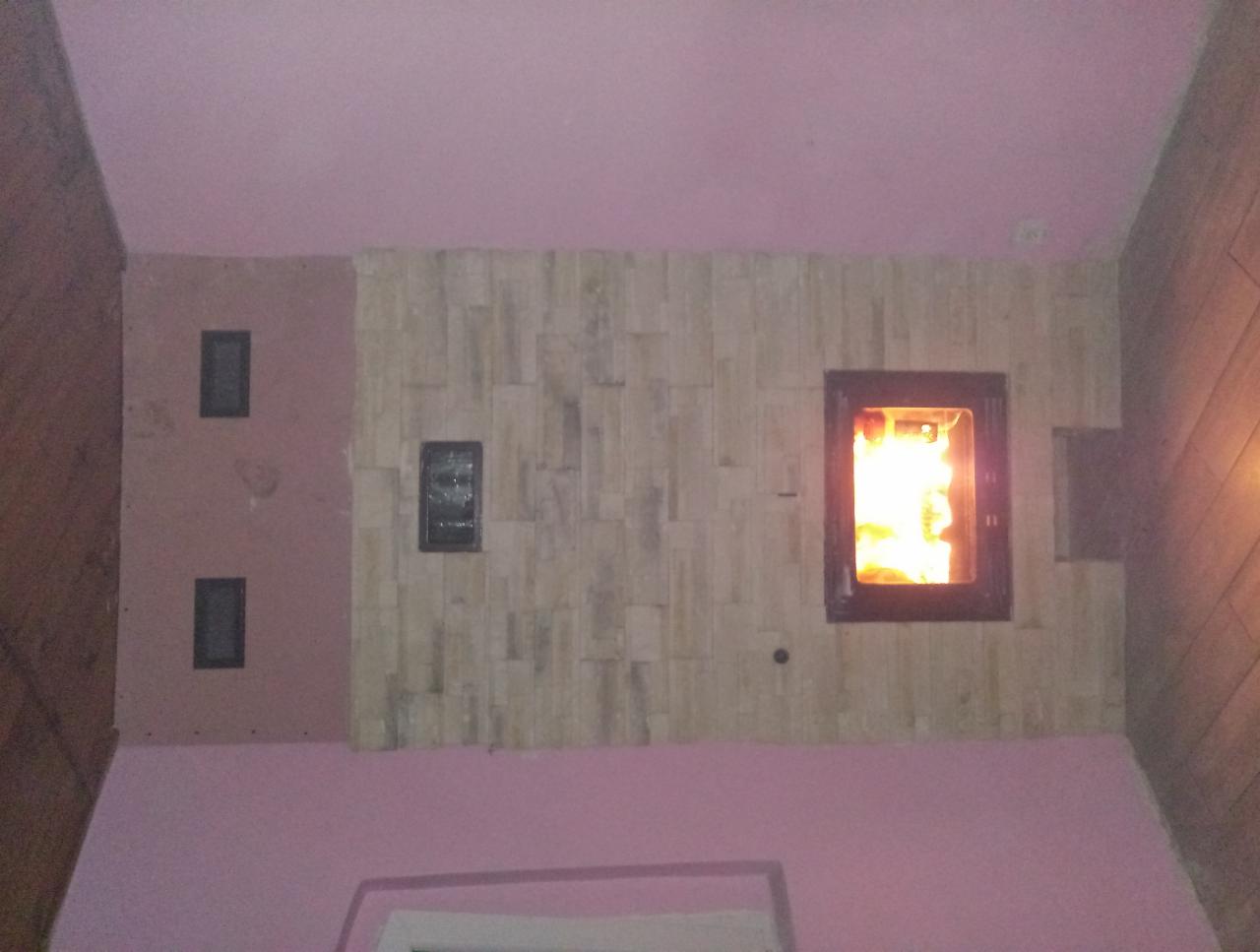 Wnętrze z wkładem kominkowym obudowanym dekoracyjną ścianą z jasnych, podłużnych płytek, widoczne płomienie w palenisku, różowe ściany i sufit.