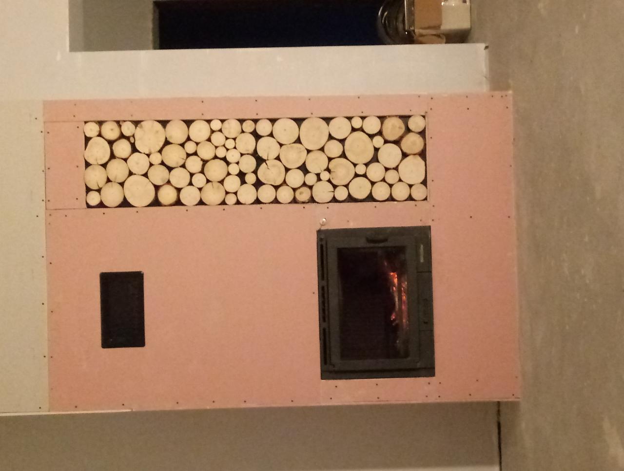 Wnętrze w trakcie montażu: obudowa z płyty gipsowej z dekoracyjnym panelem z przekrojów drewna nad wkładem kominkowym z palącym się ogniem widocznym przez szybę.