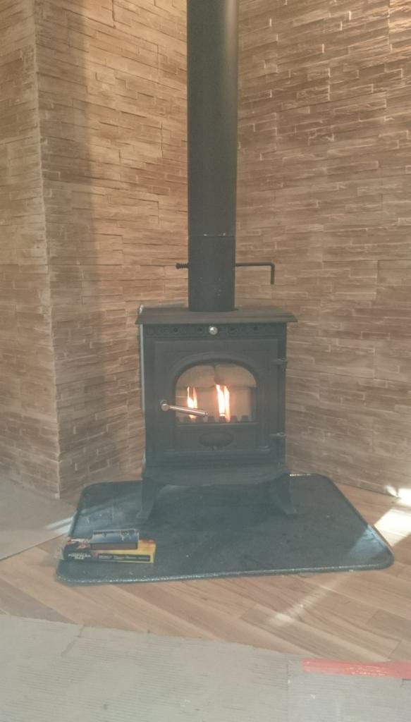 Żeliwny piec wolnostojący z płomieniami wewnątrz, ustawiony na czarnej, ochronnej macie na drewnianej podłodze, z kominem i dekoracyjną ścianą z imitacji kamienia w tle.