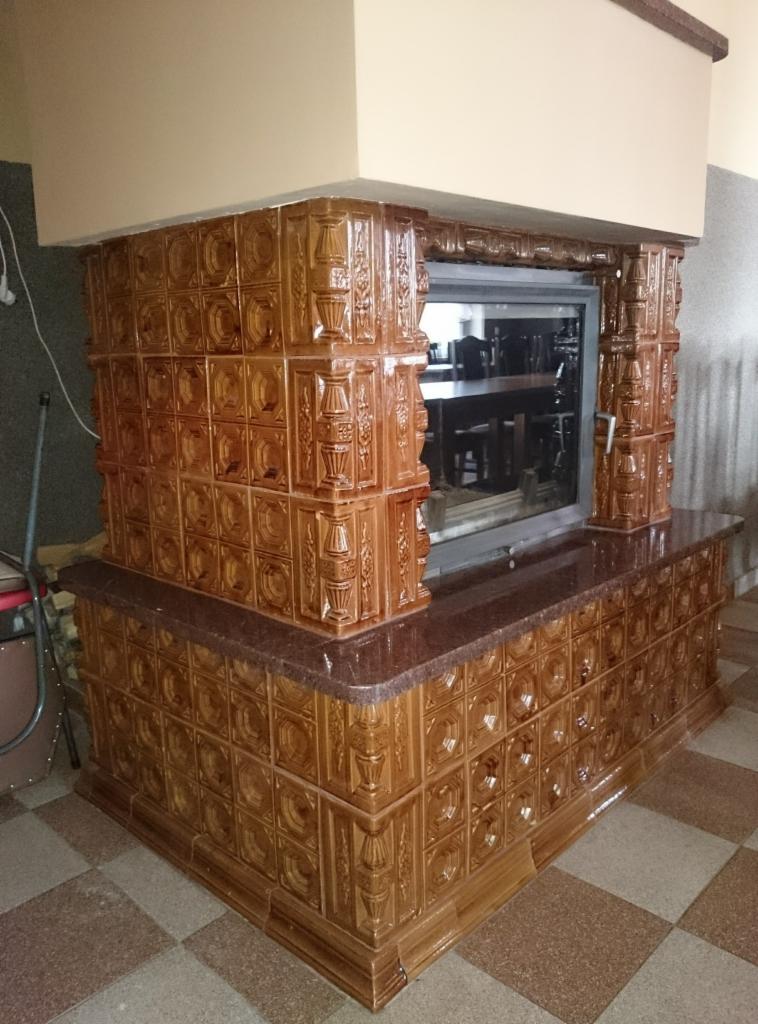 Brązowy piec kaflowy z przeszklonym paleniskiem, wykończony blatem z ciemnego kamienia, na tle brązowo-beżowej podłogi z płytek.