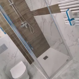 Nowoczesna kabina prysznicowa z brodzikiem, toaleta wisząca i grzejnik drabinkowy w wykończonej łazience z marmurowymi płytkami i drewnopodobnymi akcentami.