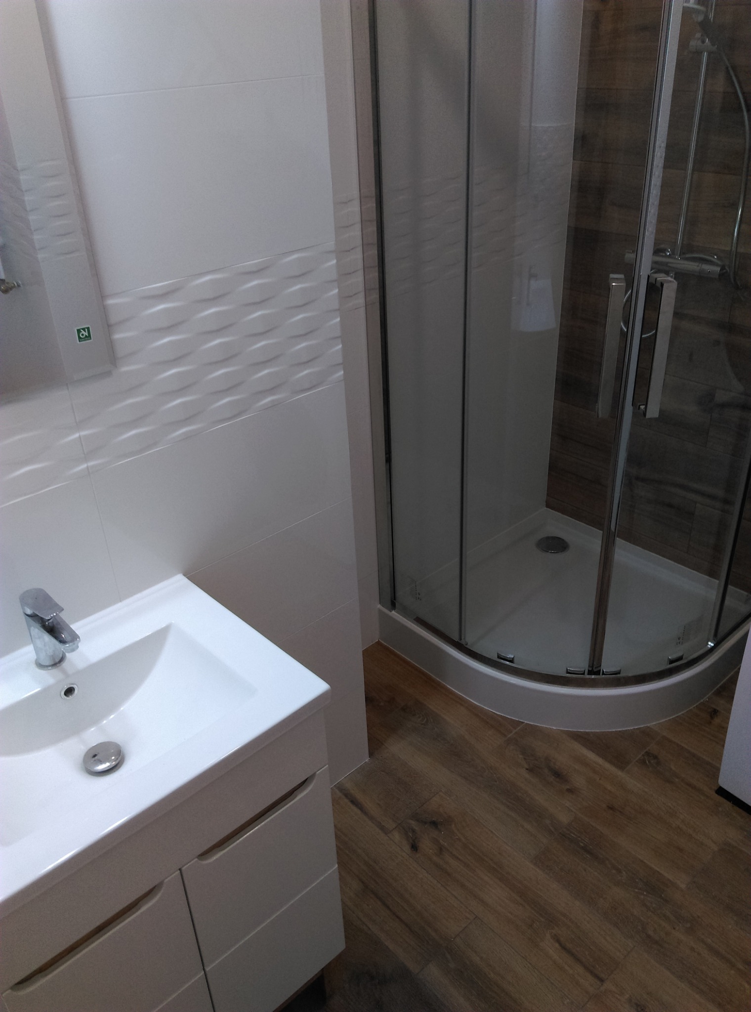 Nowoczesna łazienka z kabiną prysznicową, umywalką z szafką i białymi płytkami z trójwymiarowym wzorem oraz podłogą imitującą drewno.