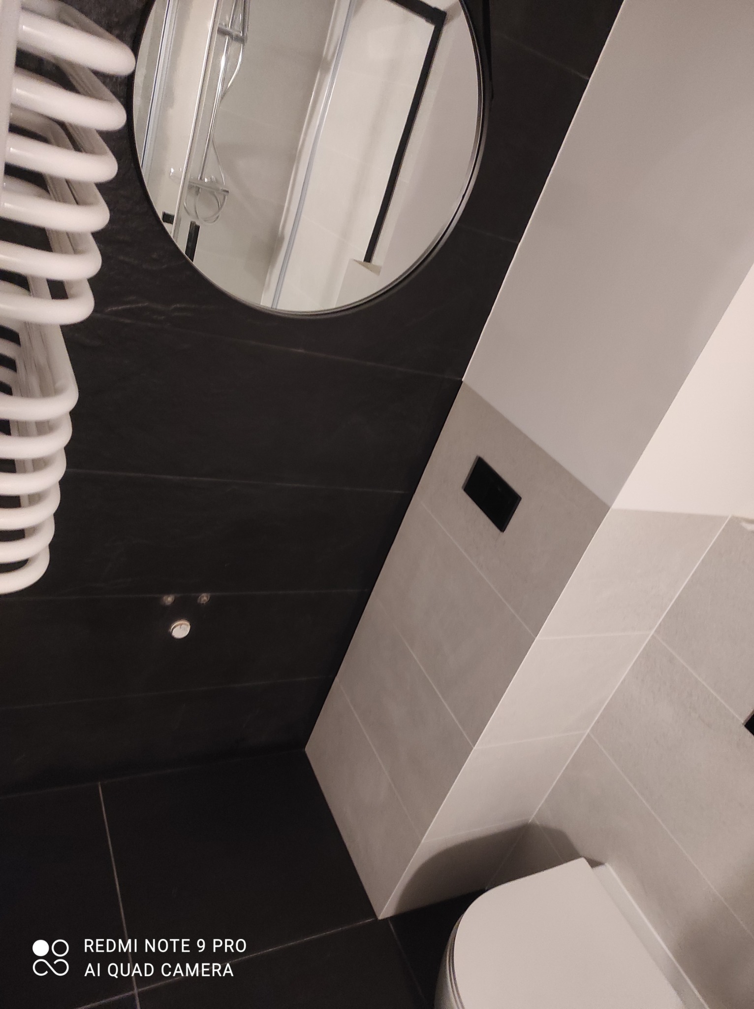 Nowoczesna łazienka z czarnymi płytkami na ścianach i podłodze, białymi płytkami na części ściany, okrągłym lustrem z czarną ramą i białym grzejnikiem drabinkowym, widziana pod kątem.