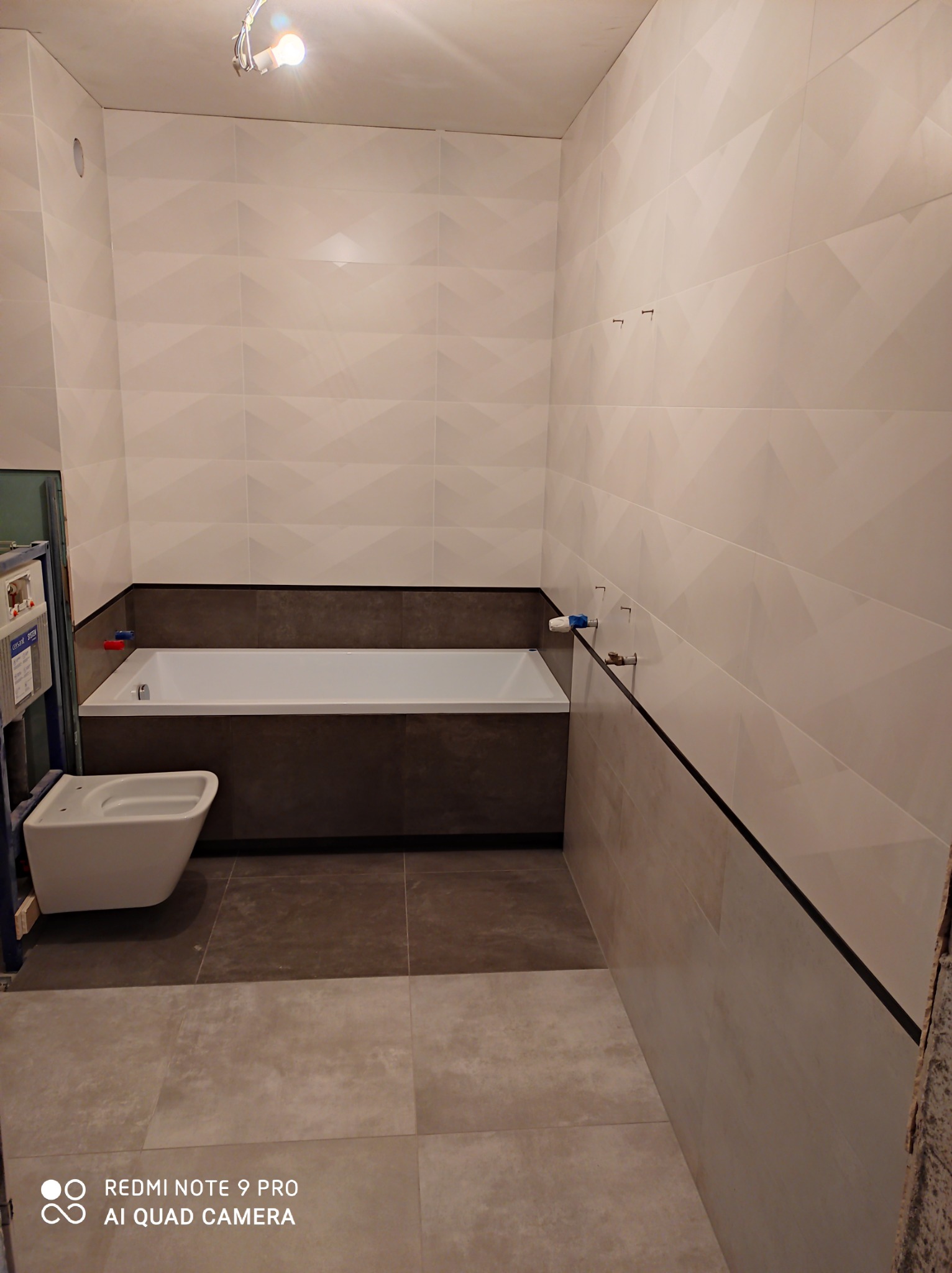 Wnętrze łazienki w trakcie prac wykończeniowych: białe płytki ścienne z geometrycznym wzorem, obudowana wanna w ciemniejszych odcieniach, częściowo zamontowana toaleta wisząca na stelażu, szare...