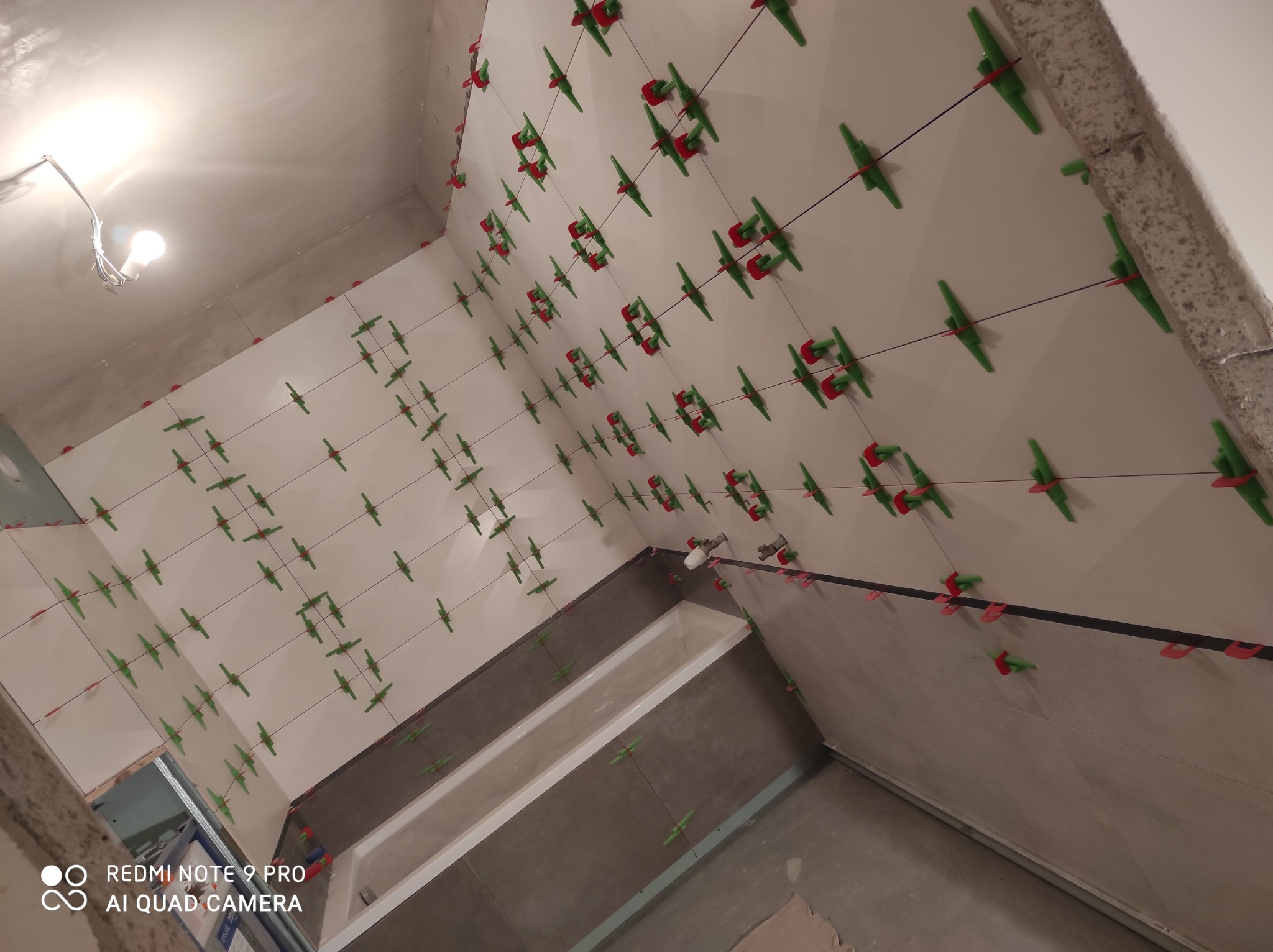 Układanie jasnych płytek na ścianach łazienki, widoczne krzyżyki dystansowe i system poziomowania płytek w kolorze zielono-czerwonym, przygotowanie do fugowania.