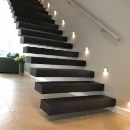 Nowoczesne, minimalistyczne schody z ciemnego drewna, oświetlone punktowymi lampami ściennymi, z białymi poręczami i podłogą w jasnym odcieniu drewna ułożoną w jodełkę.