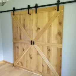 Rustykalne, przesuwne drzwi dwuskrzydłowe z jasnego drewna z czarnymi okuciami, zamontowane na szynie na tle szarej ściany i jasnej podłogi.