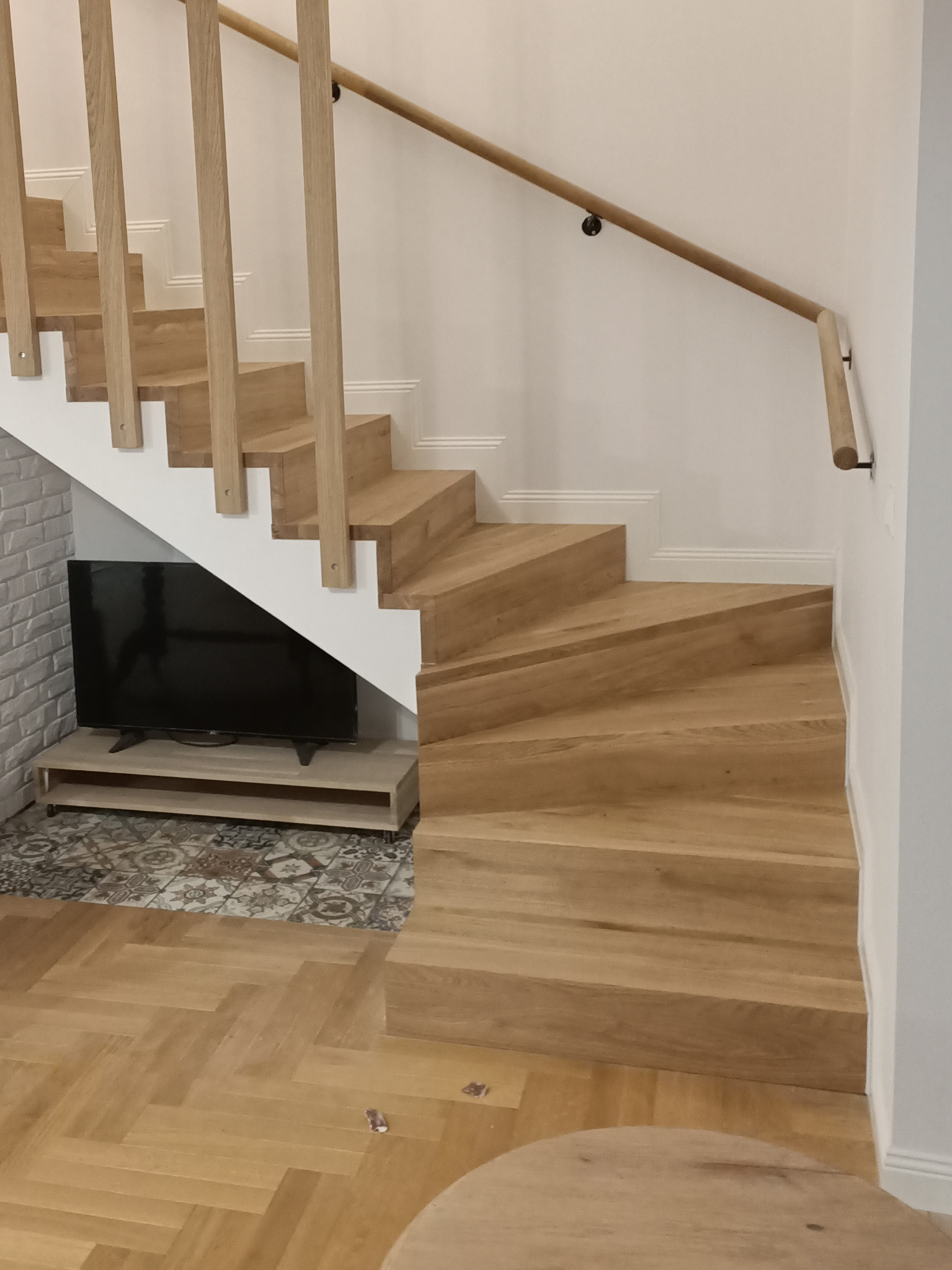 Nowoczesne, jasne schody z drewna z białymi podstopnicami, zlokalizowane w rogu pokoju z telewizorem pod biegiem schodów i parkietem w jodełkę na podłodze.