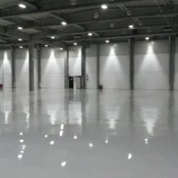 Błyszcząca, jasnoszara podłoga w hali magazynowej z widocznymi słupami i oświetleniem LED odbijającym się w powierzchni.