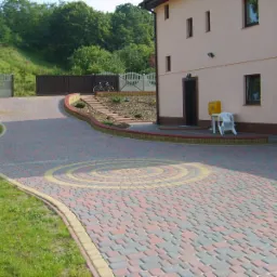 Szeroki, kręty podjazd z wielobarwnej kostki, z dekoracyjnym wzorem koła, prowadzący do domu z żółtą skrzynką pocztową i białym krzesłem.