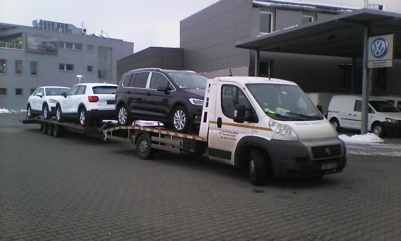 Laweta transportuje trzy samochody osobowe, dwa białe i jeden czarny, na tle budynku z logo Volkswagena w pochmurny dzień.