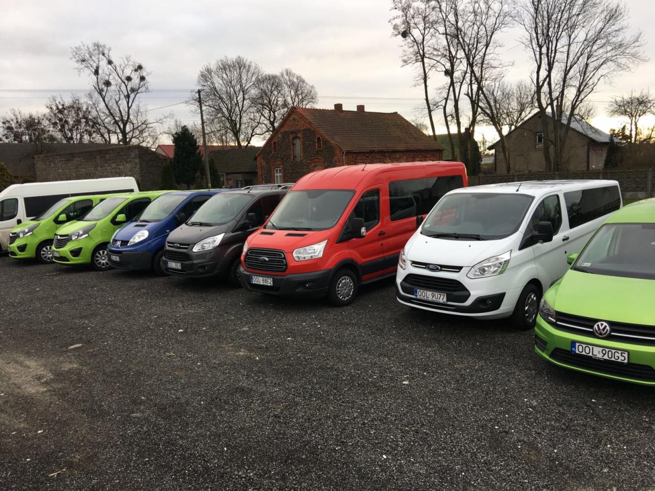 Rząd kolorowych busów marki Ford i VW na żwirowym parkingu, z budynkami w tle. Pojazdy o różnych barwach: zielony, niebieski, czerwony i biały. Pochmurne niebo.