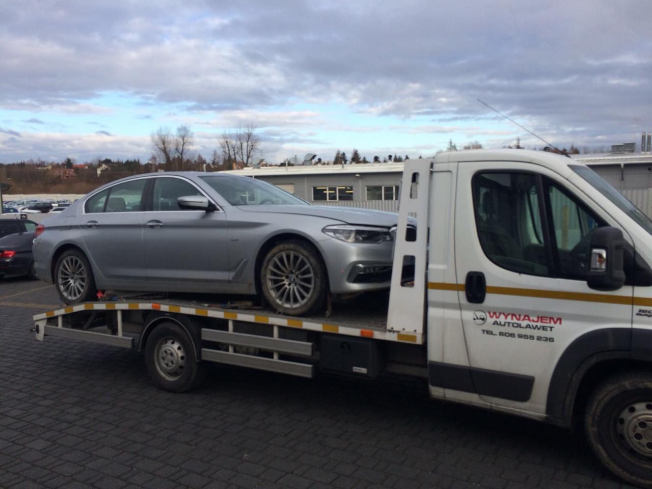 Srebrny samochód osobowy marki BMW załadowany na białą lawetę z widocznym logo firmy transportowej na boku kabiny, zaparkowaną na placu z kostki brukowej.