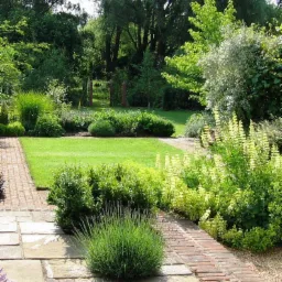 Starannie zaprojektowany ogr&oacute;d z r&oacute;wno przystrzyżonym trawnikiem, krzewami bukszpanu i ścieżką z cegły prowadzącą przez rabaty kwiatowe, zakończoną drewnianą konstrukcją w tle.