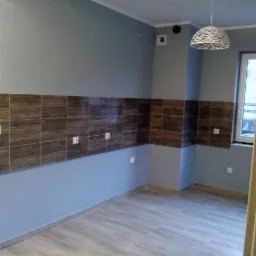 Świeżo wyremontowane pomieszczenie z brązowymi płytkami na ścianie do połowy wysokości, szarą farbą powyżej, jasnoszarą podłogą imitującą drewno i nowoczesną lampą wiszącą.