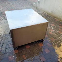 Kanciasta, metalowa skrzynia na kółkach stoi na mokrym brukowanym podłożu na zewnątrz.