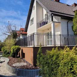 Nowoczesny dom z balkonem i tarasem zabezpieczonym czarną metalową balustradą, otoczony zadbanym ogrodem z krzewami i ścieżką.