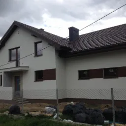 Dom jednorodzinny z ocieploną elewacją, wykończony tynkiem w kolorze złamanej bieli i z brązowymi akcentami, widoczny przez siatkę ogrodzeniową z workami śmieci.