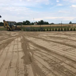 Żółta koparko-ładowarka CAT zaparkowana obok białego domu. W tle widoczne pole za ogrodzeniem z siatki i rząd małych drzewek. Na pierwszym planie wyrównany teren z widocznymi śladami opon.
