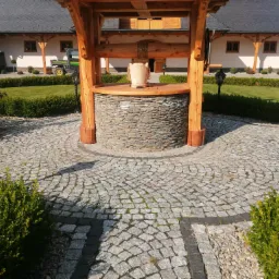 Studnia z drewnianą konstrukcją i kamienną obudową na dziedzińcu wyłożonym kostką granitową w promieniowym wzorze, w tle budynek z ciemnym dachem i starannie utrzymany trawnik.