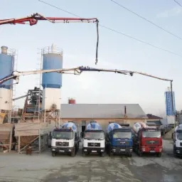 Betoniarnia z zaparkowanymi betonomieszarkami różnych kolorów, dwie z wysięgnikami do podawania betonu, budynek w tle.