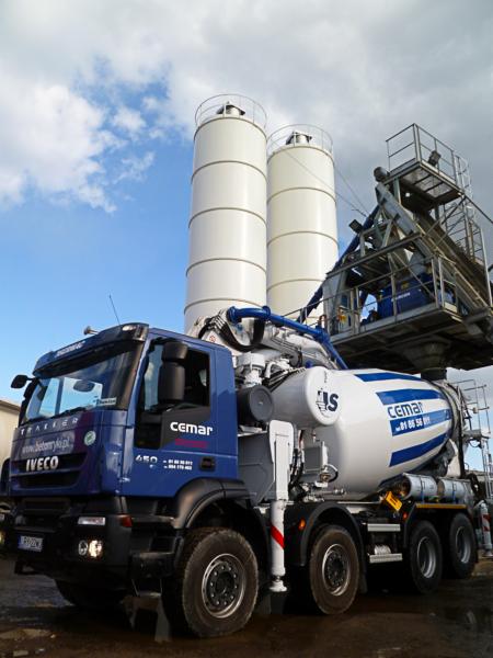 Ciężarówka betonomieszarki marki Iveco w kolorze niebieskim z białym zbiornikiem na beton, na tle silosów i pochmurnego nieba.
