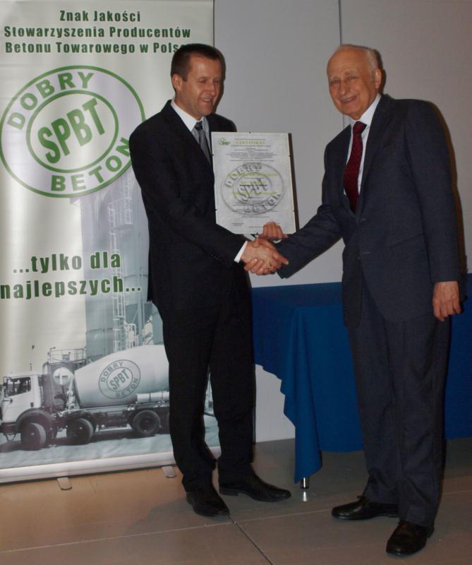 Dwóch mężczyzn w garniturach ściska sobie dłonie, trzymając certyfikat 'Dobry Beton SPBT', na tle baneru z logiem Stowarzyszenia Producentów Betonu Towarowego i zdjęciem betonomieszarki.
