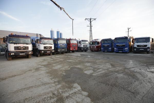 Flota betonomieszarek MAN różnych kolorów ustawiona na placu budowy z widocznymi silosami w tle i ramieniem pompy do betonu.